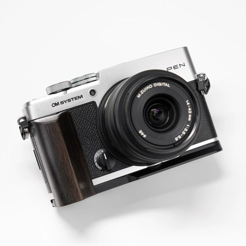 A silver and black Om System PEN camera with a dark wood-textured grip and a black Mjuico Digital lens, photographed on a clean white background.