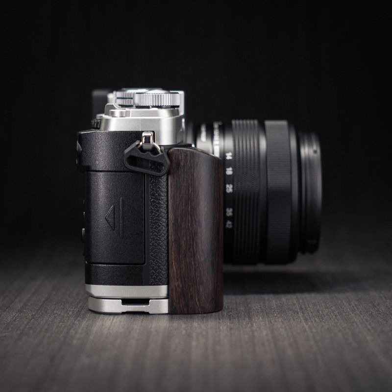 A close-up of a camera body with a dark brown wood grip on the right side, shown standing on a dark textured surface with a black lens blurred in the background. YW Design Handcrafted Wooden Grip for OM-System PEN