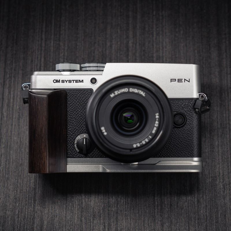 A silver and black Om System Pen camera with a dark wood grip attachment and a black lens, photographed from above on a dark textured background.