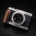 Panasonic Lumix compact camera with a silver body, black textured grip, and a brown wooden handle, shown on a dark textured background.