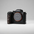 Sony Alpha 7R camera body with a black grip attachment and an orange accent ring around the lens protector, resting on a dark gray textured fabric background.