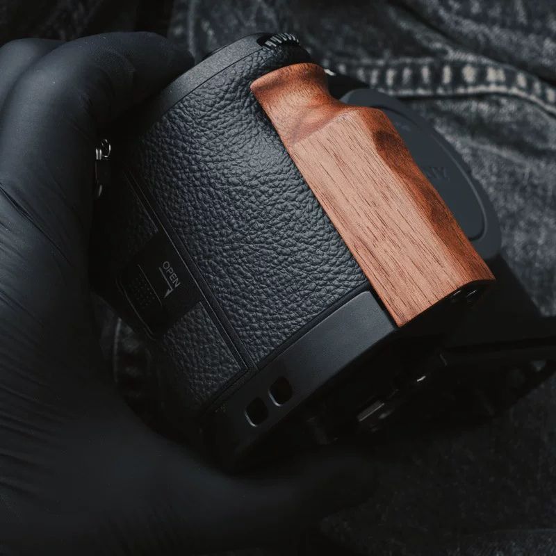 A close-up of a camera grip attachment with a textured black leather body and a warm brown wood inlay, held by a hand wearing a black glove against a dark denim background.