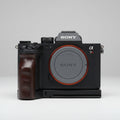Sony Alpha 7R camera body with a black grip attachment and a black lens cap with an orange ring, resting on a dark gray textured fabric background.