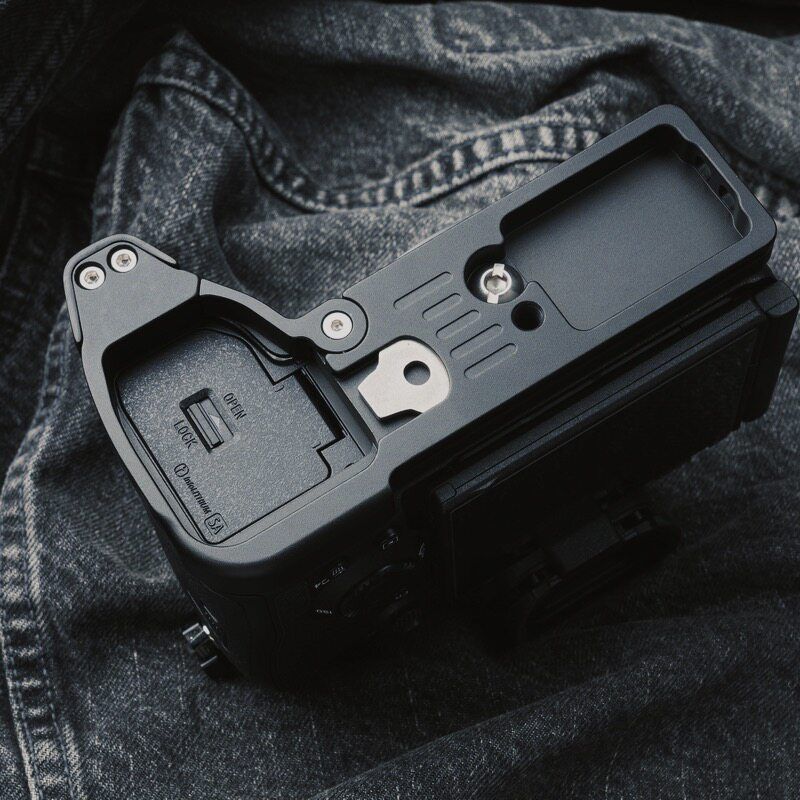 A black camera accessory cover or grip plate mounted on the top of a compact camera, photographed close-up on dark denim fabric.
